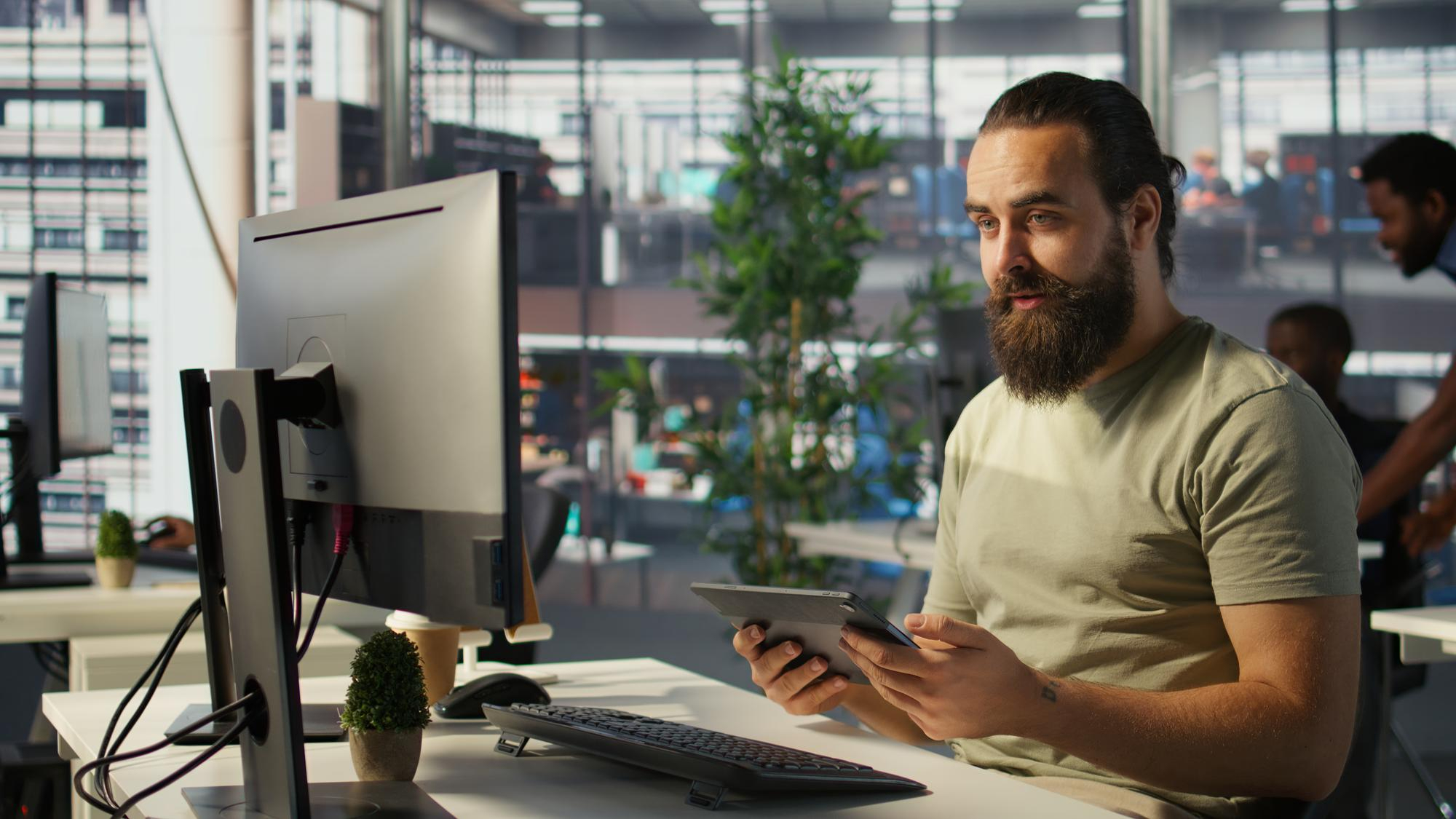 Man at a desk with a computer and tablet, holding his tablet.