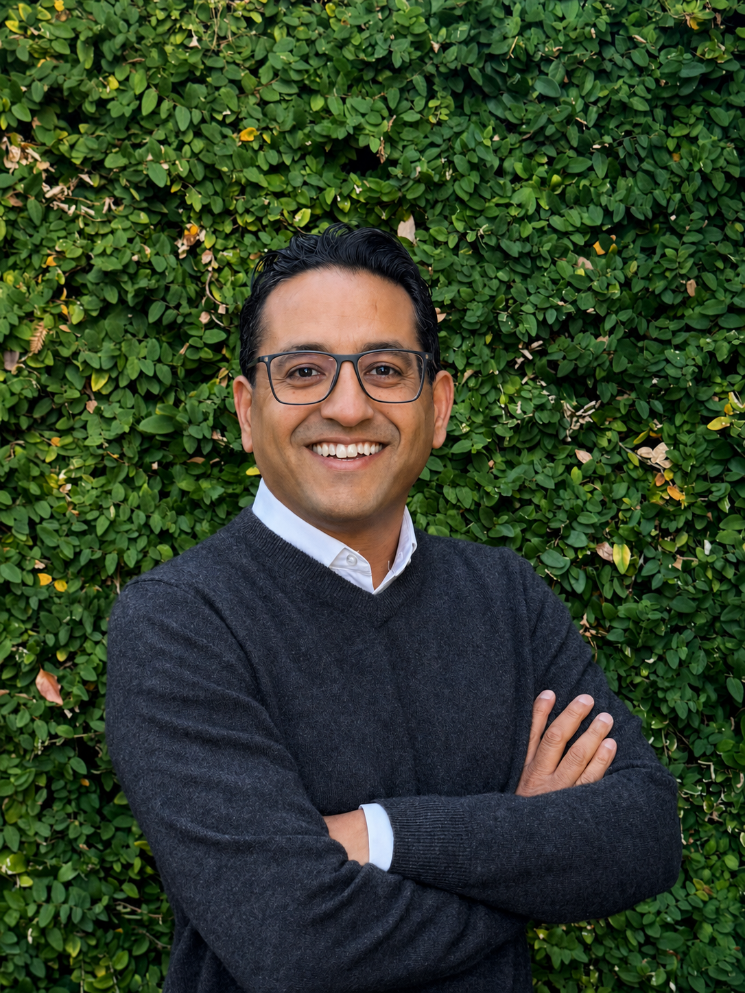 Rahul Shah with folded arms and smiling in front of green bush