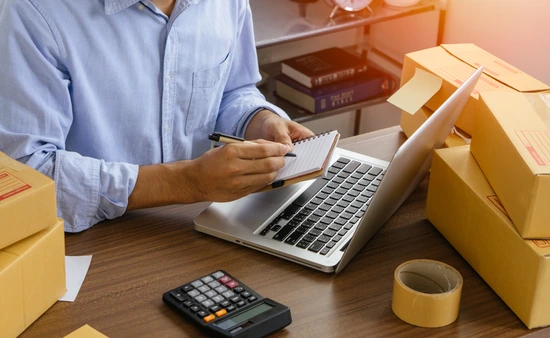 Person working at desk with laptop, calculator, and shipping boxes.