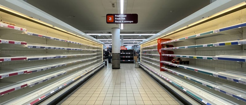 Empty store shelves with a sign indicating sections for fresh fish and bacon sausages.