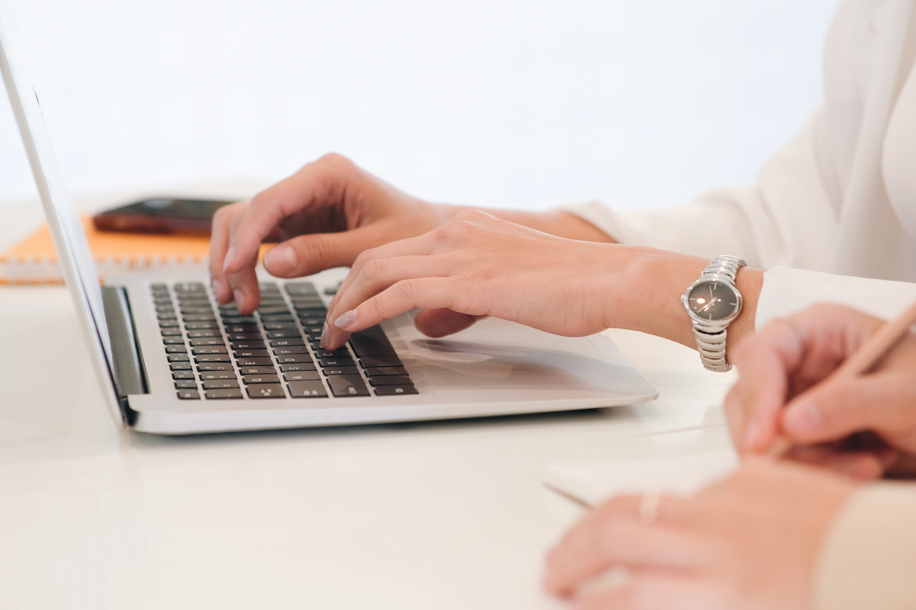 Two people working on a laptop, one typing and the other writing with a pencil.
